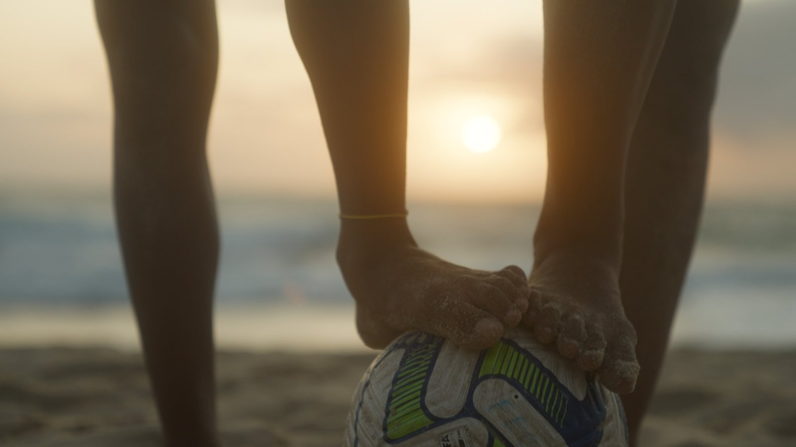 Mãos cobertas de areia apoiadas na bola, com o sol ao fundo.