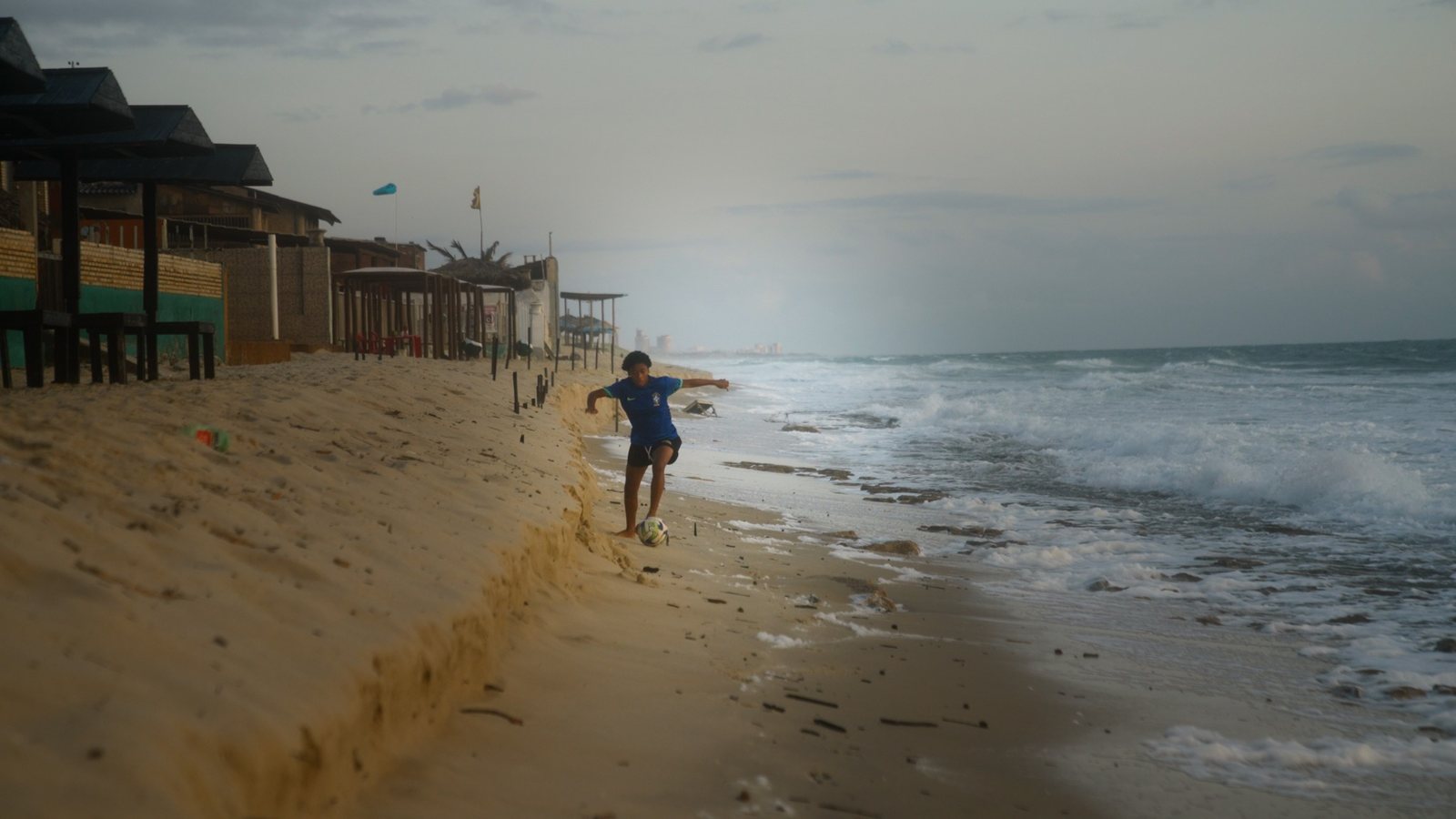 Isadora joga bola descalça na praia de Fortaleza, entre as barracas e o mar.